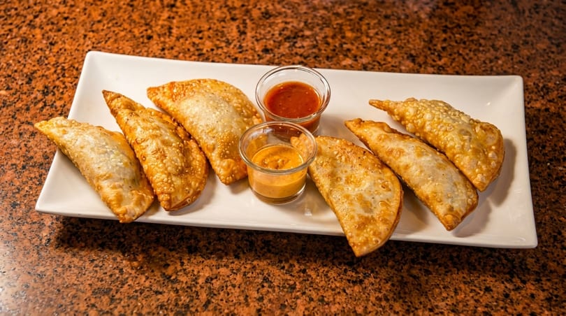 Six golden-brown empanadas and two dipping sauces on a white rectangular plate.