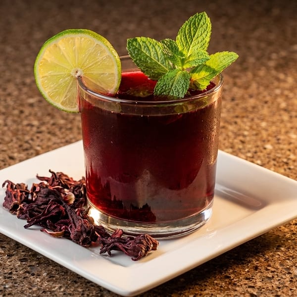 A vibrant red hibiscus drink in a glass, garnished with lime and mint, on a white plate.