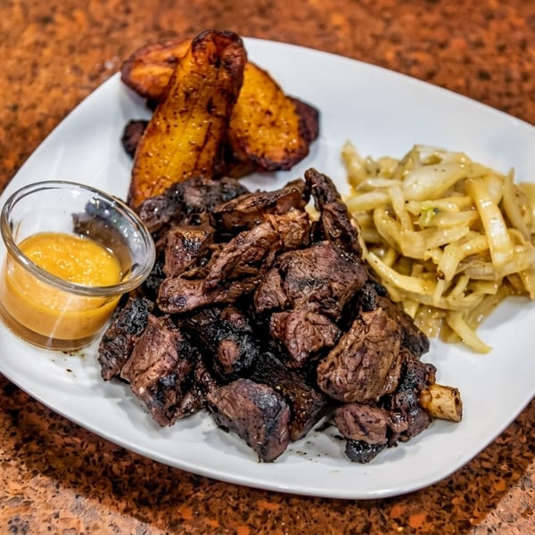 Grilled meat chunks with plantains, sautéed onions, and a dipping sauce on a white plate.