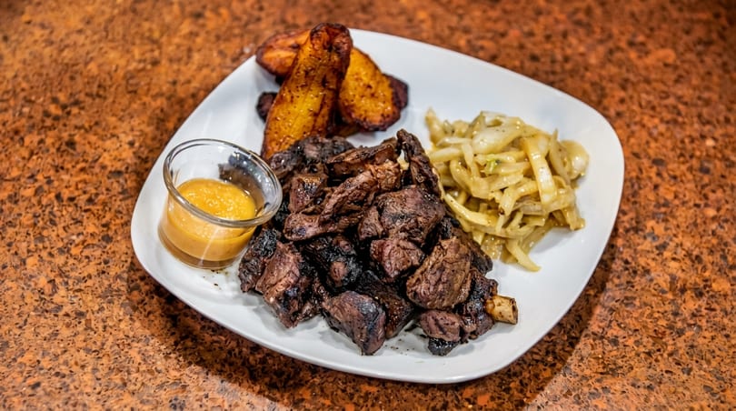 Grilled meat chunks with plantains, sautéed onions, and a dipping sauce on a white plate.