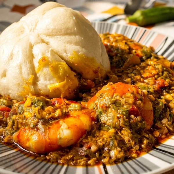 A plate of Nigerian okra soup with shrimp and a large mound of fufu on a striped plate.