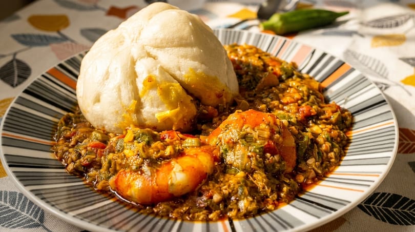 A plate of Nigerian okra soup with shrimp and a large mound of fufu on a striped plate.