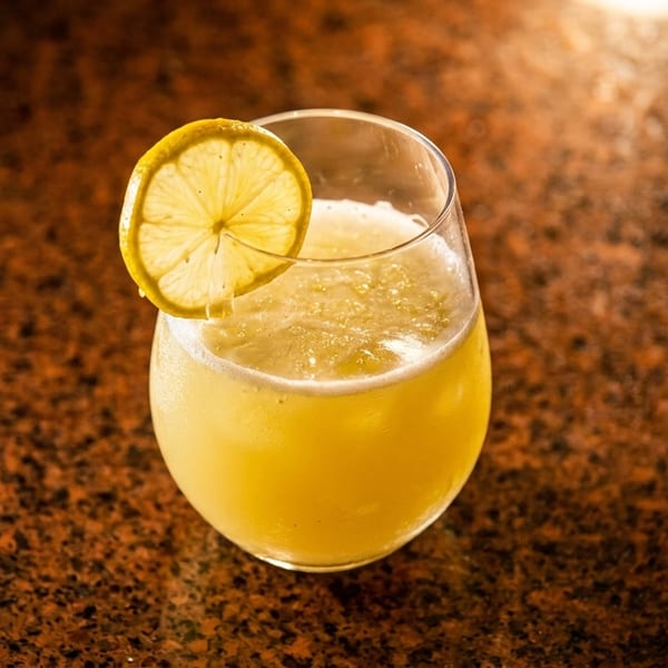 A refreshing yellow cocktail in a stemless glass, garnished with a lemon slice on a dark speckled surface.