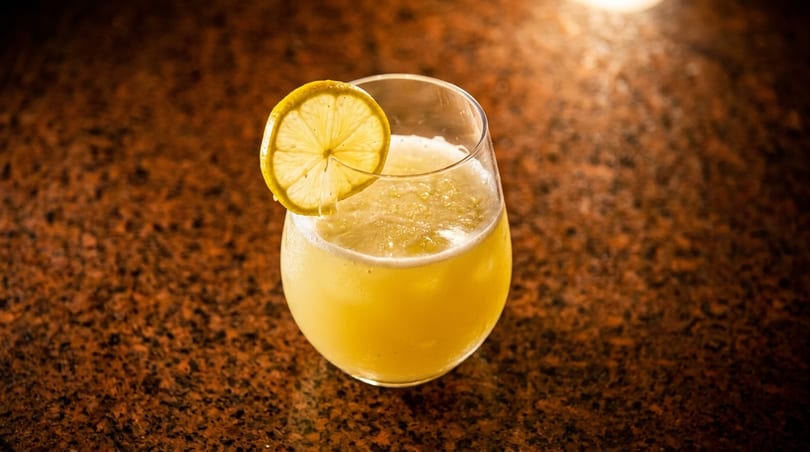 A refreshing yellow cocktail in a stemless glass, garnished with a lemon slice on a dark speckled surface.